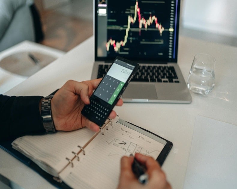 Person at a white desk uses a handheld calculator over an open notebook with diagrams; a laptop displays a stock chart in the background, and a glass of water sits nearby.