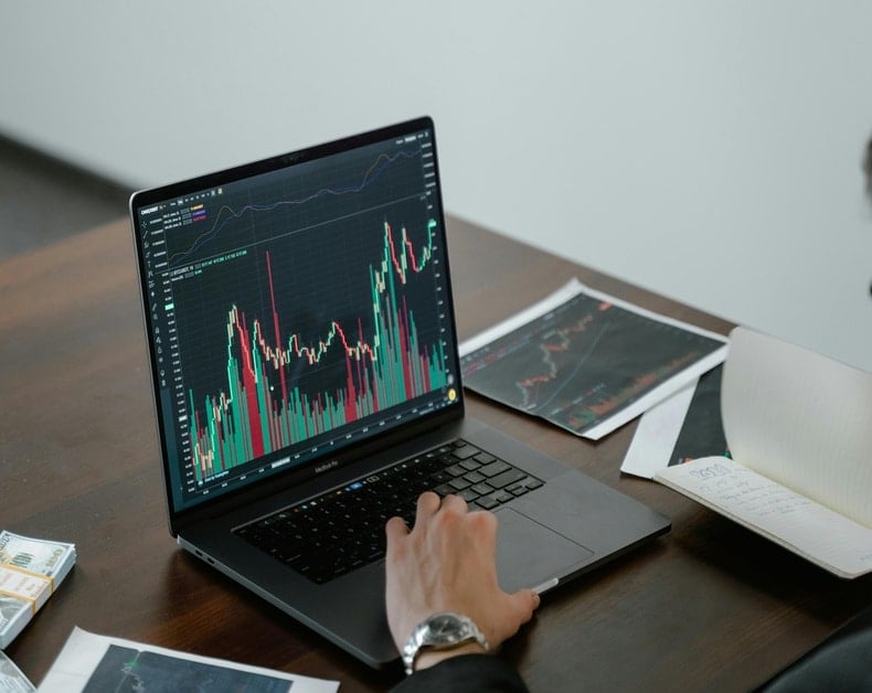 Laptop screen shows colorful stock charts and candlestick graph on a wooden desk, with papers and a wristwatch nearby.