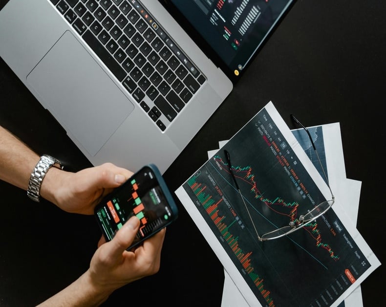 Person using a smartphone with financial trading app next to a laptop and printed stock charts on a desk.