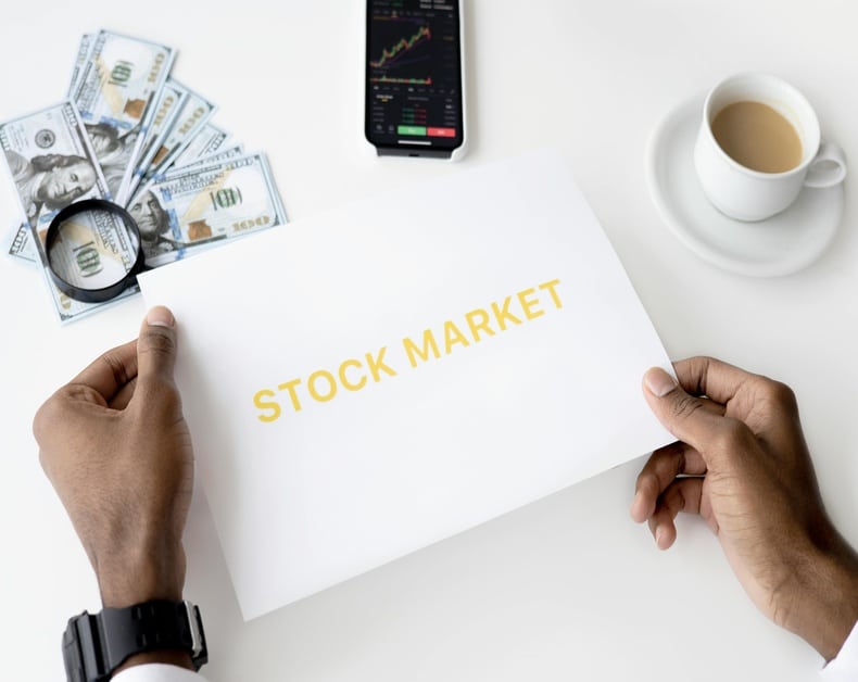 Hands hold a sheet reading 'STOCK MARKET' with a magnifying glass, cash, a smartphone showing stock charts, and a cup of coffee nearby on a white desk.
