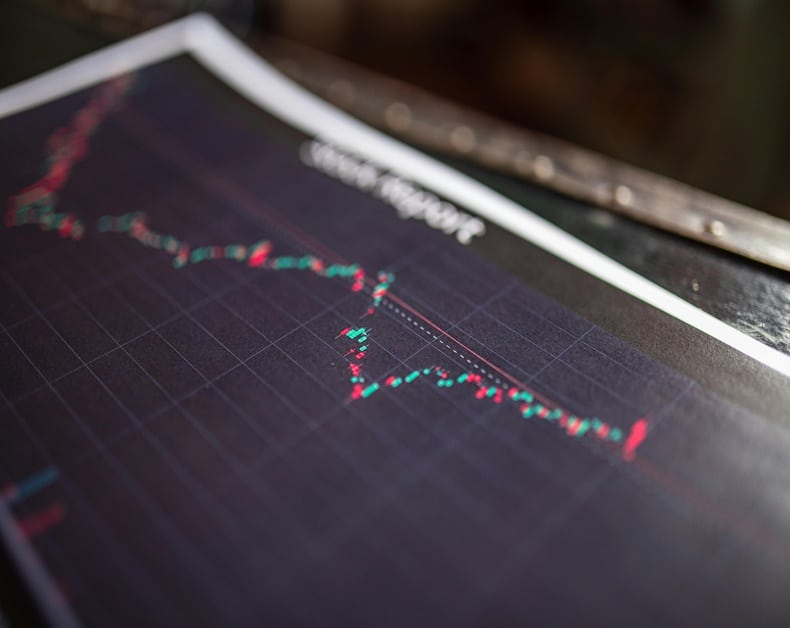 Close-up of a printed stock chart with red and green candlesticks on a grid, showing market fluctuation downwards.", "Printed financial graph displaying a fluctuating stock price with red and green markers on a dark grid.", "Stock market chart on paper illustrating a downward trend and price volatility.", "Financial chart image used to convey market fluctuations and trends."