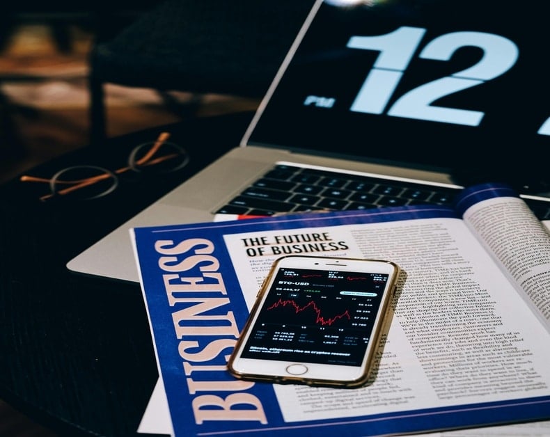 A laptop, magazine titled 'Business News', and a smartphone displaying stock charts on a desk with glasses nearby.