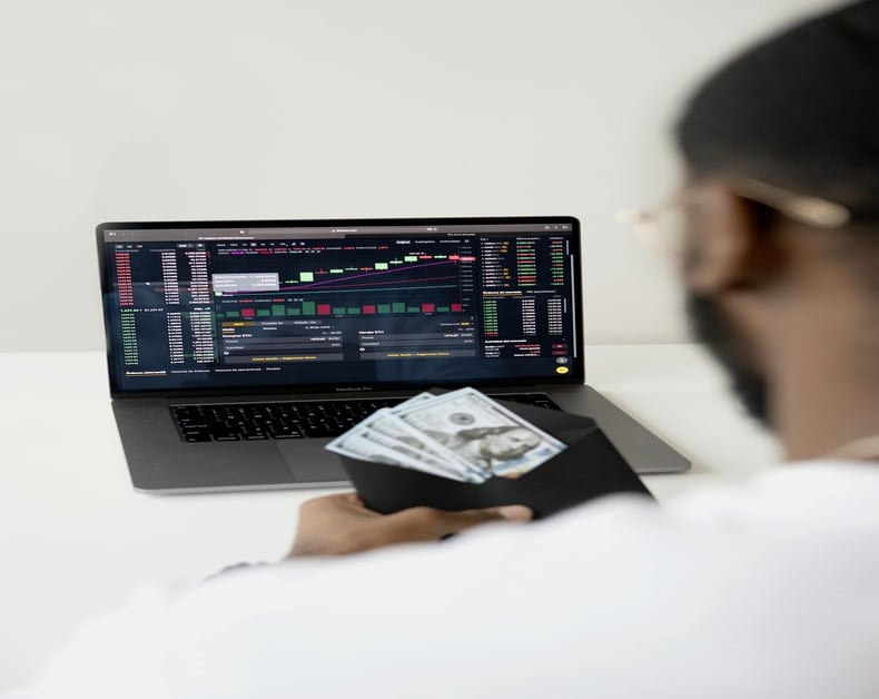 Person holding cash in a wallet while looking at a laptop screen displaying stock trading charts and data.