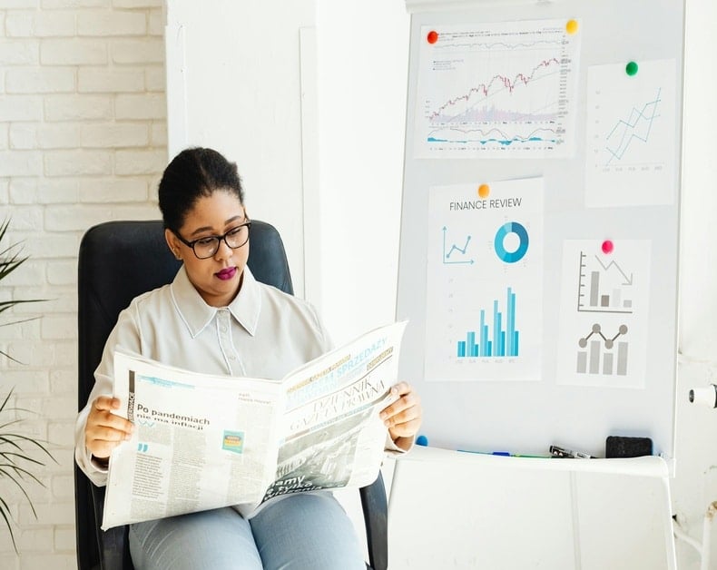 Woman in glasses sits in a black chair reading a newspaper in a bright office, with a whiteboard of finance charts nearby.