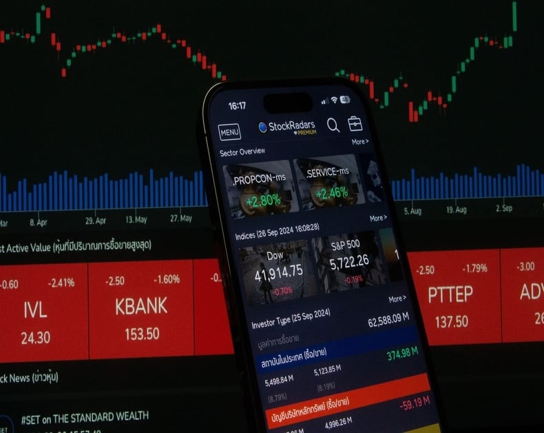 Smartphone screen showing a stock market app in front of a trading dashboard with red/green chart candles on a dark background.