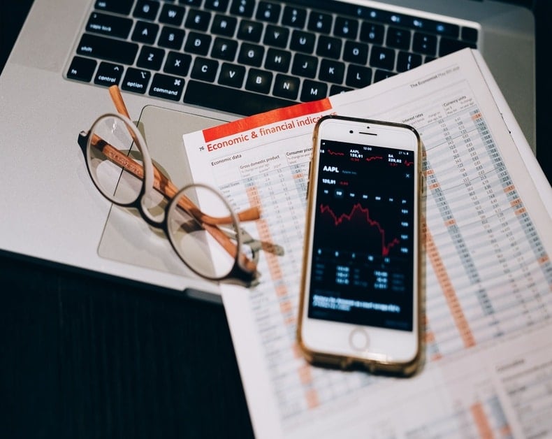 Glasses rest on a laptop with financial documents and a smartphone showing stock charts or market data.