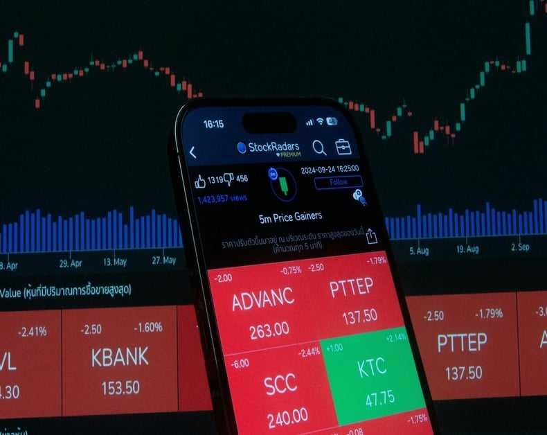 Close-up of a smartphone screen showing a stock trading app with red and green price tiles, over a dark market chart background.