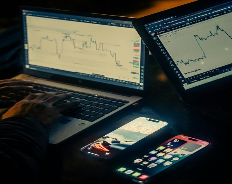 Two laptops open to stock charts (one with candlesticks, the other with a rising line) and three smartphones on a dark desk, a trading setup.