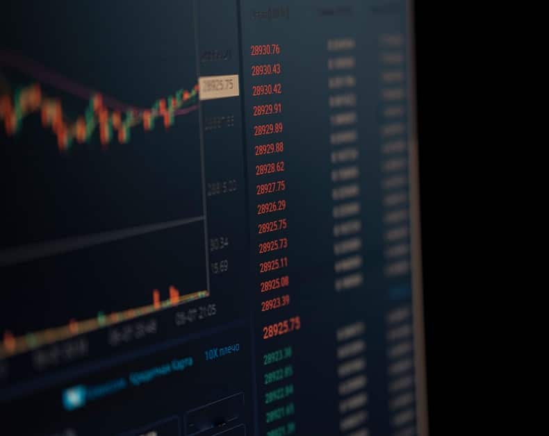 Close-up of a monitor displaying stock market data with red price numbers and a blurred candlestick chart on a dark background.