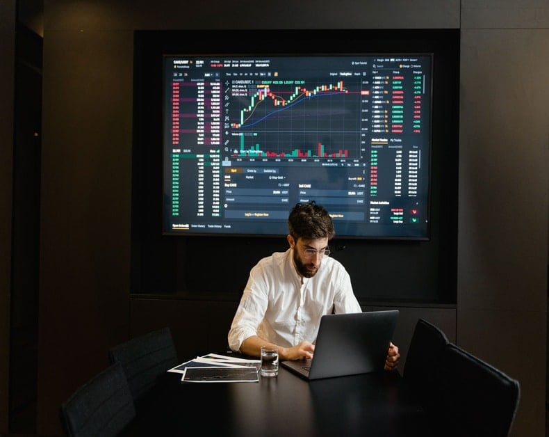 Man in a white shirt typing on a laptop at a dark conference room, a large stock market dashboard with charts and data glowing on the wall behind him