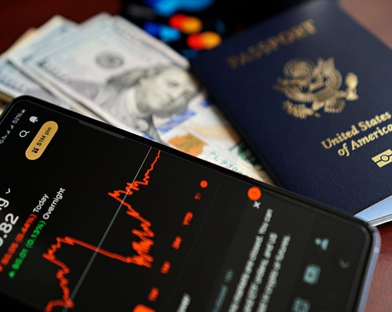 Close-up of a smartphone displaying a red stock market graph, with a U.S. passport and scattered cash in the background.