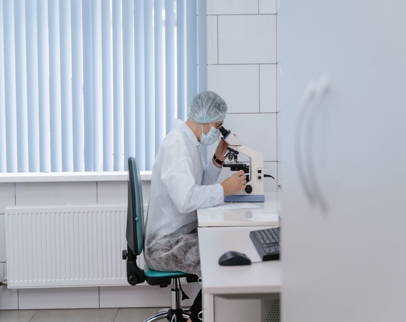 Scientist in a lab coat and hairnet using a microscope at a clean bench, wearing a mask in a medical lab.