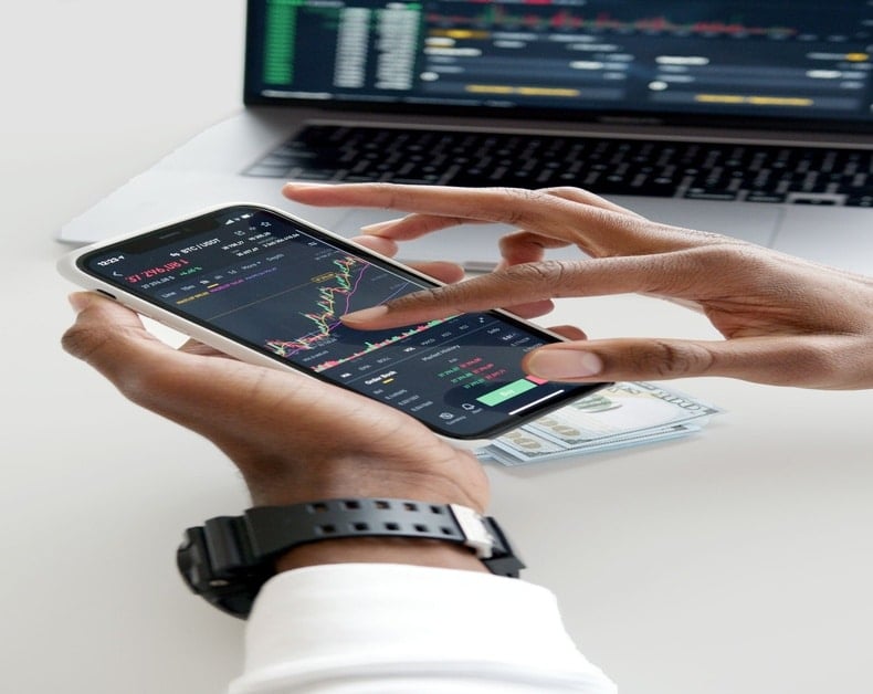 Person holding a smartphone showing stock trading charts, with a laptop and cash on the desk in the background.