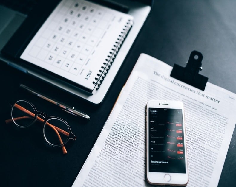 Desk scene with glasses, pen, a spiral notebook, calendar, and a smartphone displaying stock quotes on top of a newspaper about digital currencies.