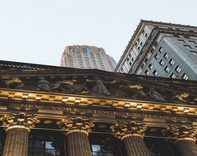 Façade of the New York Stock Exchange with ornate columns and sculpted figures, set against tall surrounding buildings.