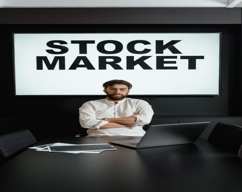 Man with folded arms seated at a dark conference table in front of a large screen that reads STOCK MARKET.