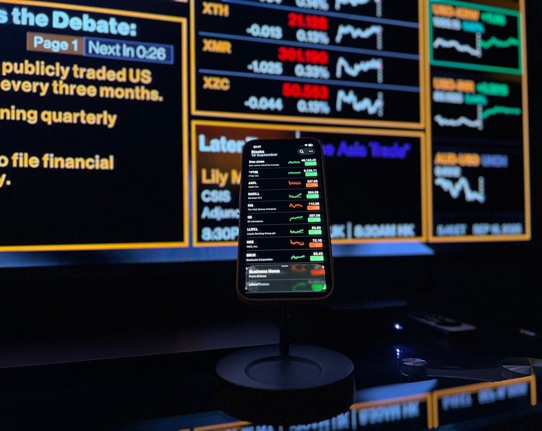 Smartphone on a stand in front of a wall of stock ticker boards displaying live quotes in green and red.