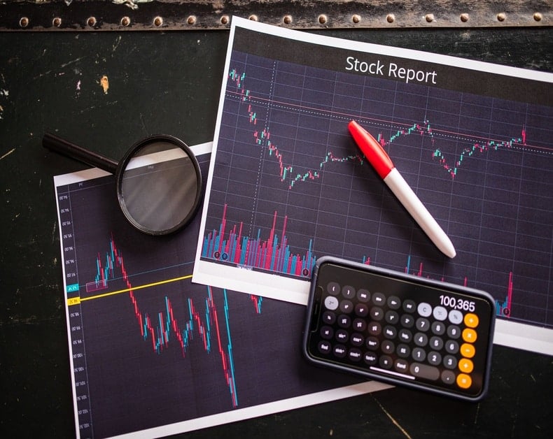 Stock market charts on printed reports with a magnifying glass, a red-and-white marker, and a calculator.