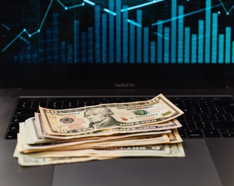 Stack of US dollar bills on a laptop keyboard with a blue stock chart on screen, conveying finance and trading
